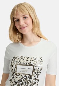 Hvid bomulds T-shirt med korte ærmer, der har et leopardmønster i guld og sort, samt et "BE YOURSELF N°34" label.