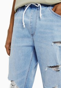 Närbild av ljusblå slitna jeansshorts med fransade hål och ett vitt snöre knutet i midjan, bärs av en person med händerna i fickorna.