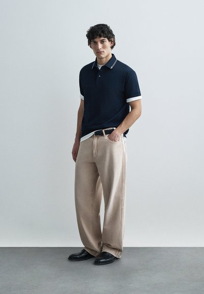 Ναυτικό πόλο με λευκό γιακά και μανσέτες, συνδυασμένο με ανοιχτό μπεζ φαρδιά παντελόνια και μαύρα παπούτσια. Μινιμαλιστικός σχεδιασμός και ουδέτερα χρώματα.