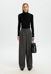 Zwarte lange mouwen top met een nekstrik, gecombineerd met hoge taille grijze wijde broek. Houdt een kleine zwarte gestructureerde handtas vast.