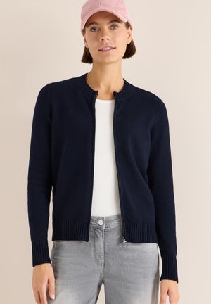 Cardigan zip-up blu navy realizzato in morbido tessuto, con polsini e orlo a costine, indossato sopra una maglietta bianca, abbinato a jeans grigio chiaro.