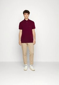 Bordeauxrotes Poloshirt mit Kragen und kurzen Ärmeln, kombiniert mit beigefarbenen, schmal geschnittenen Hosen und weißen Turnschuhen. Minimalistisches Design ohne Muster.