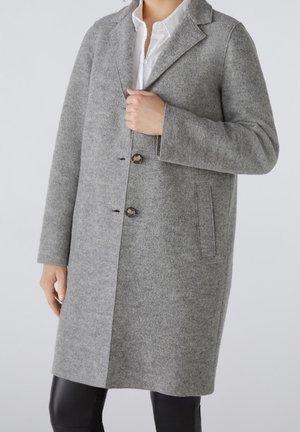 Personne portant un manteau en laine gris longueur genou avec deux boutons, un col cranté, des poches latérales, et une chemise blanche en dessous.