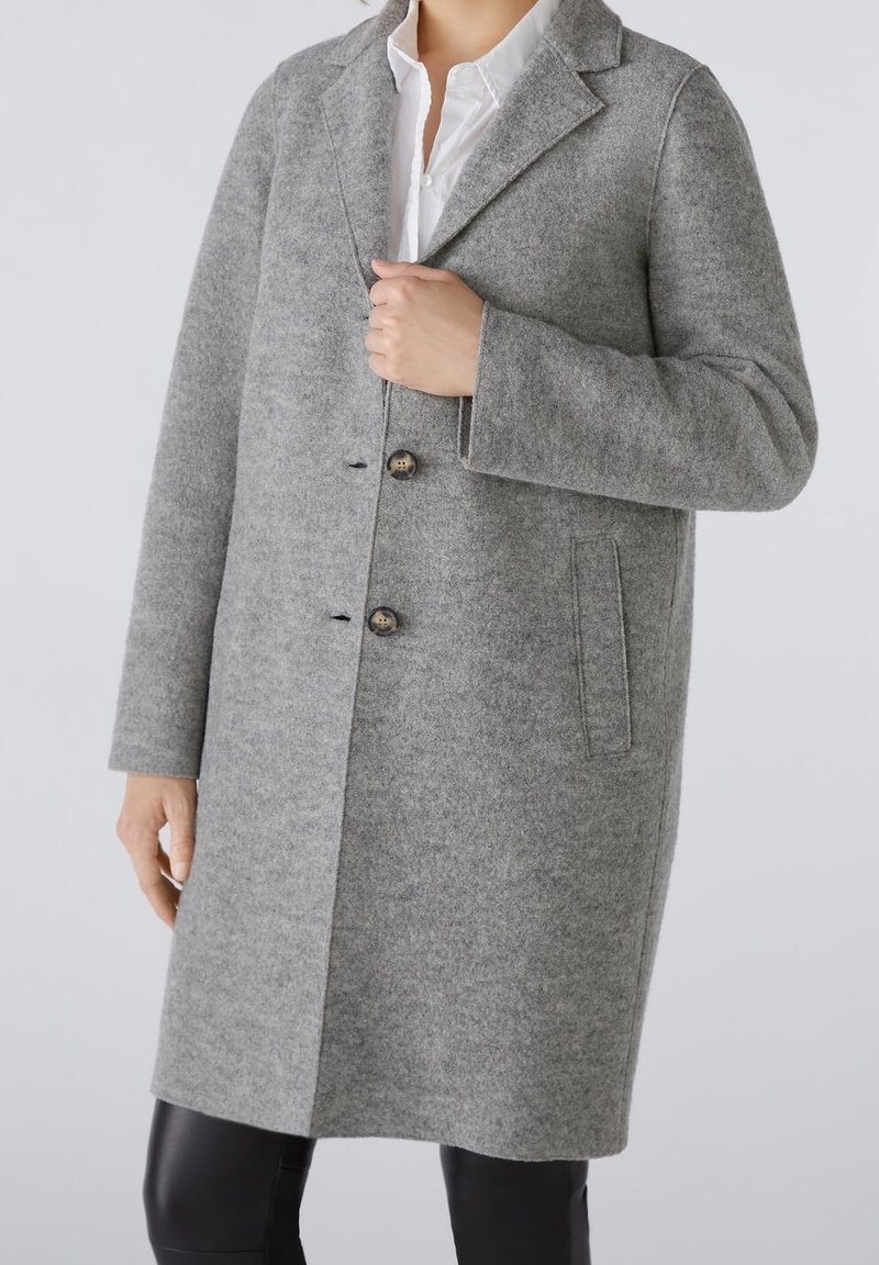 Personne portant un manteau en laine gris longueur genou avec deux boutons, un col cranté, des poches latérales, et une chemise blanche en dessous.