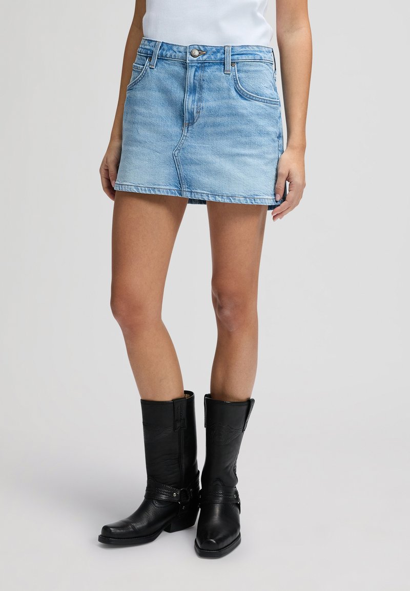 Γυναίκα που φοράει ανοιχτό μπλε τζιν μίνι φούστα, λευκό τοπ και μαύρες δερμάτινες μπότες μέχρι τη μέση της γάμπας, σε όρθια θέση μπροστά σε απλό φόντο.