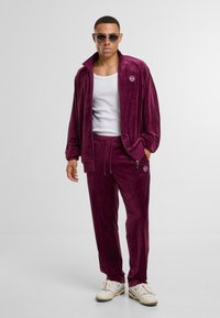 Pluszowy, burgundowy dres z zamkiem błyskawicznym w kurtce i spodniami na sznurku, w połączeniu z białym topem na ramiączkach, podkreślony okrągłymi okularami przeciwsłonecznymi.