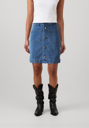 Femme portant une jupe en jean bleue boutonnée avec des poches avant, un haut blanc sans manches et des bottes noires souples mi-mollet sur un fond gris.