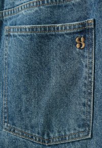 Poche arrière d'un jean en denim bleu avec des coutures marron et un petit logo marron brodé dans le coin supérieur droit.