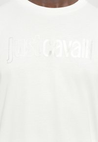 Balta T-krekls ar spīdīgu sudraba drukāto tekstu "justcavalli" dažādos fontu svaros. Mīksta, gluda auduma tekstūra.