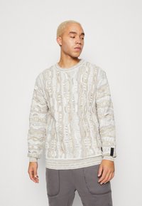 Knit sweater in white and beige with textured cable pattern, ribbed cuffs and hem, and logo accents. Casual fit with round neckline.