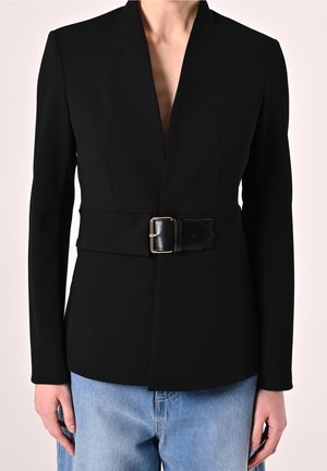 Blazer noir sur mesure avec large ceinture et boucle dorée, porté avec un jean en denim bleu clair, vu du cou aux hanches, les mains reposant le long du corps.