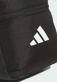 Černý texturovaný batoh s výrazným bílým logem adidas a černým zipem. Materiál vypadá odolně a voděodolně.