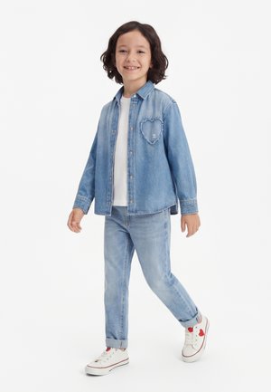 Smaidīgs bērns iet, ģērbies gaiši zilā džinsa jaciņā ar sirds uzšuvumu, baltā krekliņā, atbilstošās džinsās un baltās sporta kurpēs ar sarkanu sirds detaļu.