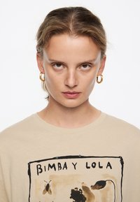 Beige puuvillane T-särk, millel on graafiline trükk tumedate kontuuridega, kujutades lille- ja putukate motiive, väikeste mustade aktsentidega disainis.