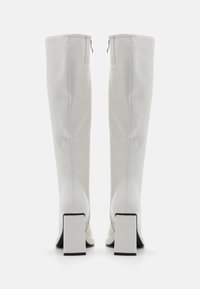 Bottes blanches montant jusqu'au genou en matière synthétique lisse. Talons carrés avec accents noirs. Fermeture zippée à l'arrière avec détails de couture minimalistes.