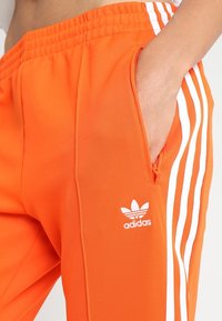 Oranssit urheiluhousut, joissa on valkoinen Adidas-logo ja kolme valkoista raitaa sivuilla, sisältävät elastisen vyötärön ja sivutaskun.