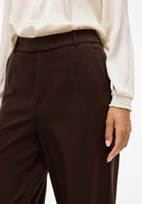 Pantaloni marroni su misura con una texture liscia, dotati di cuciture laterali e parte frontale piatta. Abb Binati a una blusa bianca ampia.