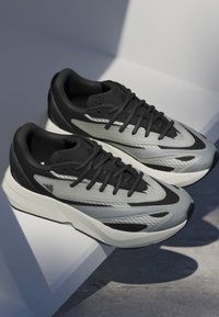 Čierne a šedé športové topánky s textúrovaným zvrškom, štíhlym dizajnom a výraznou svetlou medzipodrážkou. Obsahujú šnúrky a detail loga.