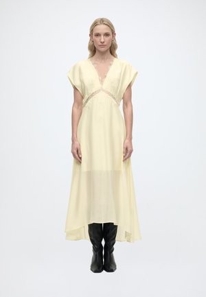 Femme blonde debout, portant une robe jaune pâle à col en V avec des détails en dentelle et des bottes noires montantes, sur un fond blanc uni.