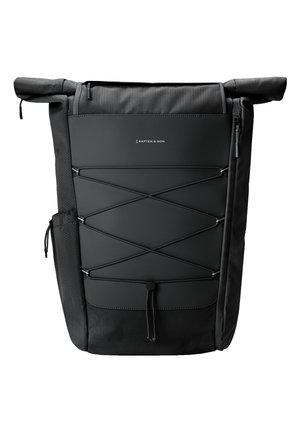Personne portant un sac à dos noir à rabat enroulable avec cordon ajustable et poches latérales, vêtue d'une veste violet clair sur un fond uni.