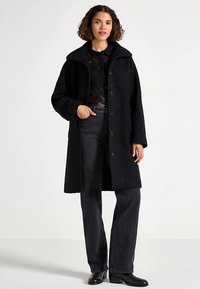 Schwarzer, flauschiger Mantel aus strukturiertem Stoff mit großem Kragen, Knopfverschluss und Seitentaschen, kombiniert mit schwarzen Jeans und Stiefeletten.