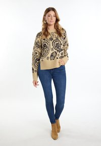 Beżowy sweter z dzianiny z czarnym wzorem paisley, ściągaczami przy mankietach i u dołu. Połączony z ciemnoniebieskimi dżinsami i jasnobrązowymi botkami za kostkę.