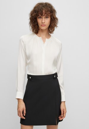 Weißes Blusenshirt mit sanftem Glanz, ausgestattet mit einer Knopfverschluss und gerafftem Ausschnitt, kombiniert mit einem schwarzen Rock mit seitlichem Schlitz und dekorativen Knöpfen.