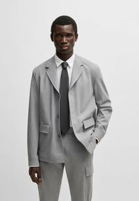 Grau maßgeschneiderter Anzug mit einem einreihigen Blazer mit aufgesetzten Taschen, kombiniert mit maßgeschneiderten Hosen. Strukturiertes Material, klassisches weißes Hemd und Krawatte.