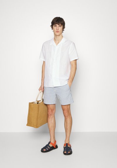 Cameriera bianca a maniche corte, pantaloni corti a righe blu chiaro, sandali neri e arancioni, mentre tiene in mano una borsa in paglia beige. Design semplice e casual.