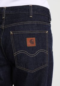 Dark blue denim jeans featuring a leather logo patch on the back pocket, contrasting yellow stitching, and a classic five-pocket design.