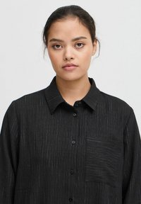 Zwarte shirt met een verticale streepjespatroon, knoopsluiting aan de voorkant en een zakje. Heeft een kraag en een gestructureerde, geweven stof.