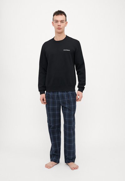 Sweatshirt noir à col rond avec lettrage blanc, associé à un pantalon de pyjama à carreaux bleu marine avec des lignes bleu clair. Le modèle est pieds nus.