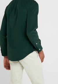 Ciemnozielona koszula z teksturowanej tkaniny, z długimi rękawami i zapięciem na guziki przy mankietach, włożona do jasnobeżowych spodni z zaokrąglonym dołem.