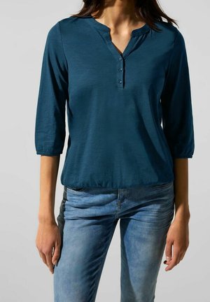 Femme portant un chemisier à manches longues couleur sarcelle avec trois boutons et un jean bleu délavé, debout devant un fond clair uni.