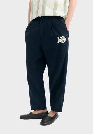 Osoba koja nosi široke crne hlače s bijelim izvezenim motivom ribe na gornjem dijelu bedra i crne cipele koje se navlače.