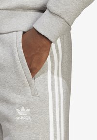 Sive trenirke s belim tridelnim dizajnom, z bočnim žepom in vezenim logotipom na levi stegn. Gladek tekstilni material.