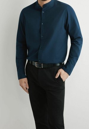 Homme portant une chemise bleu foncé à col mandarin rentrée dans un pantalon noir avec une ceinture noire, une main dans la poche, sur un fond uni.