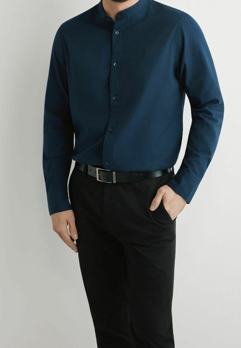 Homme portant une chemise bleu foncé à col mandarin rentrée dans un pantalon noir avec une ceinture noire, une main dans la poche, sur un fond uni.