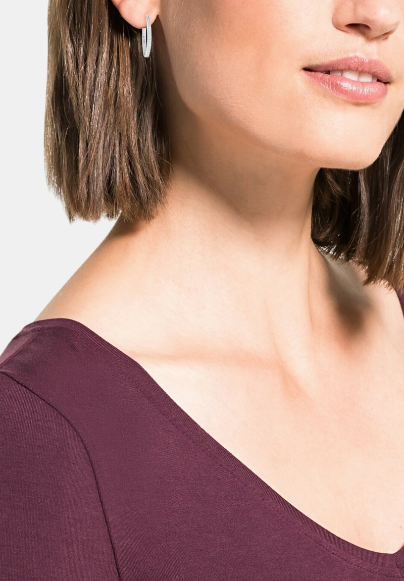 Orecchino ad anello in argento con una forma ovale stretta e una superficie leggermente testurizzata. Indossato con una maglia di un profondo bordeaux con un'ampia scollatura.