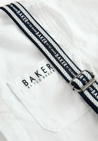 Bela tkanina s teksturiranim, tkanim pasom, ki ima mornarsko modre in bele črte. Na oznaki je napis "BAKER by TED BAKER" v črni barvi. Podrobnost s srebrnim okrasjem.