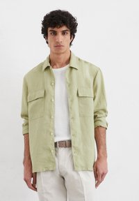 Chemise vert clair à manches longues avec deux poches poitrine, fermeture à boutons et coupe décontractée. Portée par-dessus un t-shirt blanc et associée à un pantalon clair.