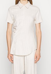 Donna che indossa una camicia a maniche corte con bottoni e strisce sottili, caratterizzata da un dettaglio arricciato laterale, abbinata a pantaloni chiari in tonalità coordinata.