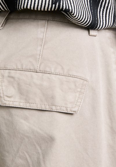 Pantalons en coton beige avec une poche arrière ; la texture du tissu est lisse avec un tissage diagonal subtil ; superposés avec une chemise rayée noire et blanche.