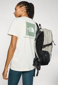 Mujer con cabello trenzado, vistiendo una camiseta blanca de The North Face y leggings en color azul verdoso, llevando una mochila gris y negra sobre un hombro.