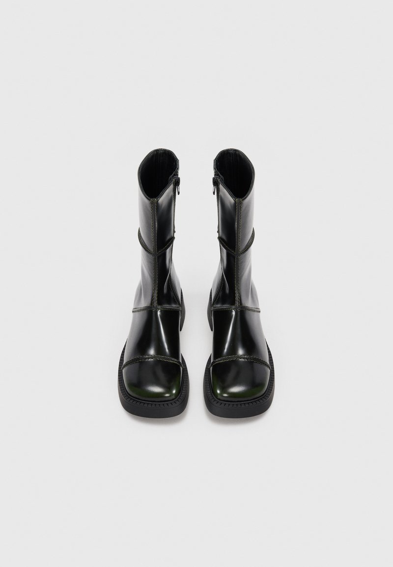 Stivali alla caviglia in pelle nera con finitura lucida, punta quadrata, zip laterali e cuciture decorative. Suola piatta in gomma con grip testurizzato.