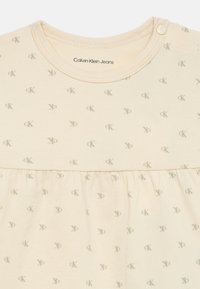 T-shirt de algodão bege com manga curta, apresentando um padrão sutil de logotipos CK repetidos. Tem um decote redondo e uma cintura franzida.