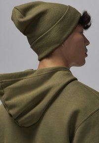 Berretto lavorato a maglia verde oliva indossato con una felpa con cappuccio verde oliva. Il tessuto appare morbido con dettagli di cucitura visibili e una vestibilità comoda.