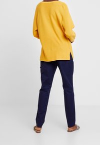 Yellow ribbed pullover with a relaxed fit, side slits; paired with navy blue slim-fit pants and tan flat sandals with snake print accents.