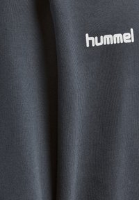 Tmavě šedá dlouhá tričko s dlouhým rukávem vyrobené z hladké látky, s bílým logem "hummel" na přední straně. Textura je měkká a žebrovaná.