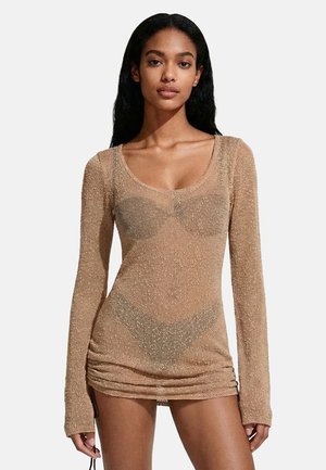 Donna con capelli lunghi e neri che indossa un mini abito beige a maniche lunghe e trasparente sopra biancheria intima coordinata, su uno sfondo semplice.